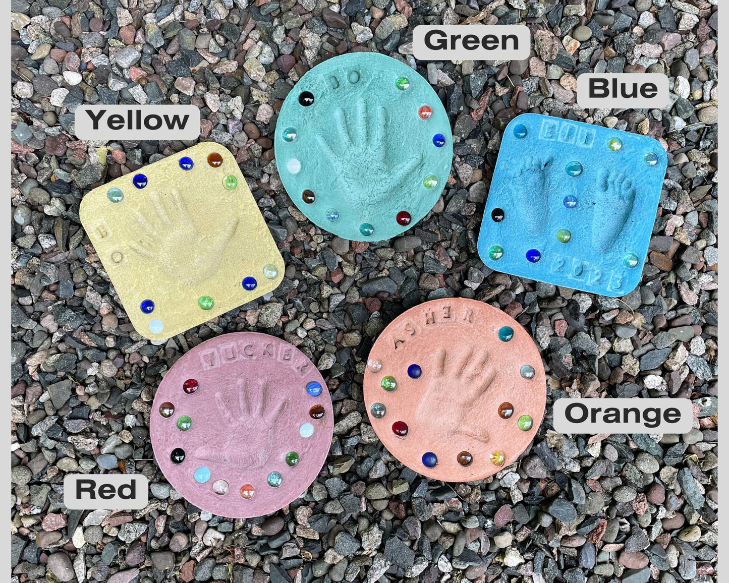 Assortment of colored stepping stones displayed on ground showcasing different designs and colors. Shows the larger sized stones with three hand prints on one stone.  Red, Blue, Yellow, Orange, Green and Grey color options shown in picture. Mama B's