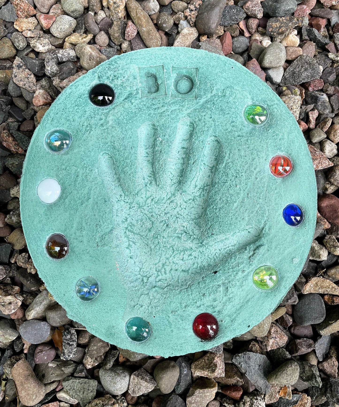 Assortment of colored stepping stones displayed on ground showcasing different designs and colors. Shows the larger sized stones with three hand prints on one stone. Red, Blue, Yellow, Orange, Green and Grey color options shown in picture. Mama B's