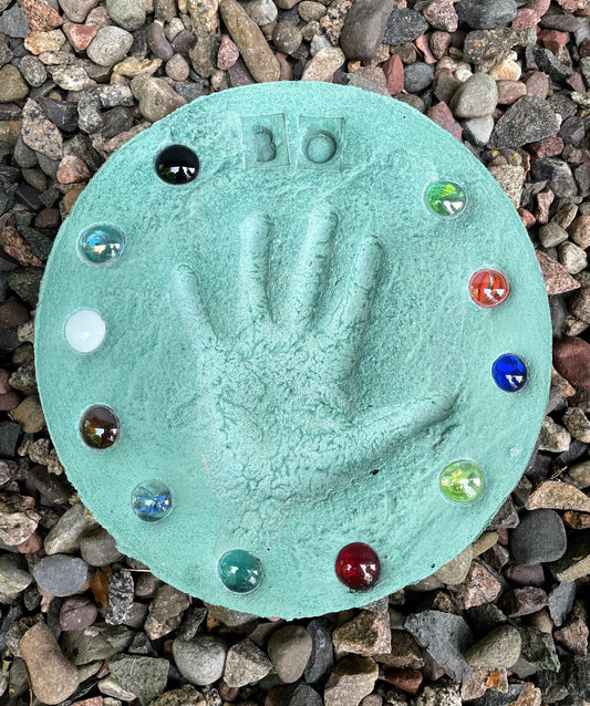 Assortment of colored stepping stones displayed on ground showcasing different designs and colors. Shows the larger sized stones with three hand prints on one stone.  Red, Blue, Yellow, Orange, Green and Grey color options shown in picture. Mama B's 